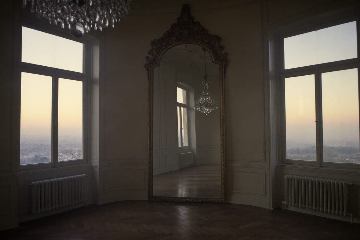 A crystal chandelier reflected in a gilt-framed mirror in an empty ballroom at dawn
