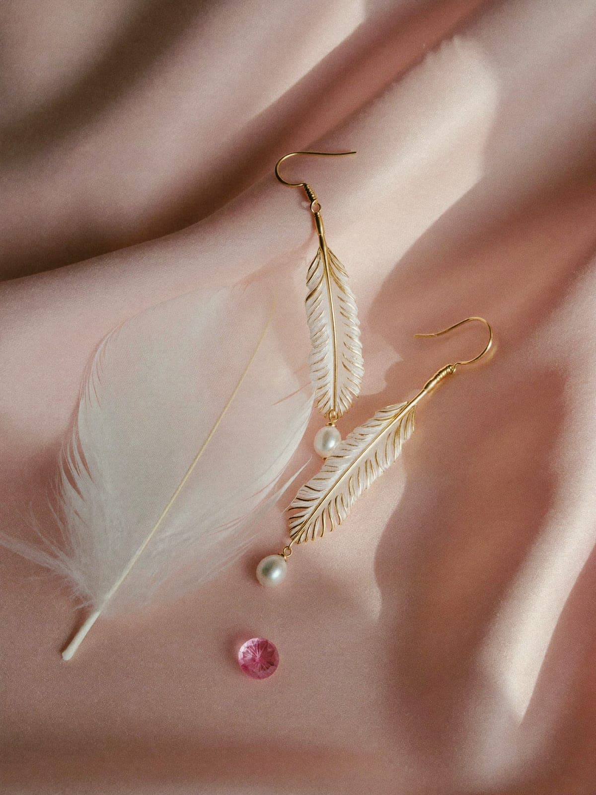 Slender gold drop earrings shaped like feathers with small pearls, beside a white feather